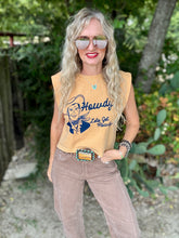 A woman wearing a sleeveless tan graphic t-shirt with a cowboy illustration and the text 'Howdy Lets Get Rowdy with brown pants, standing outdoors in front of a garden background.