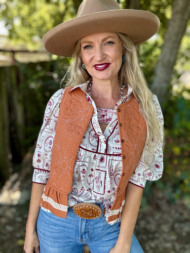 Woman wearing a cowboy hat, patterned shirt, vest, and jeans outdoors.