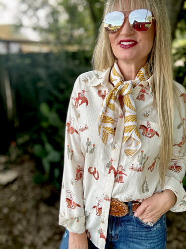 Woman wearing a patterned blouse with a scarf, sunglasses, and a belt buckle outdoors.