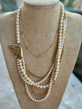 A multi-layered pearl and gold necklace displayed on a beige fabric jewelry bust. The necklace features multiple strands of white faux pearls, gold chain links, a gold leaf-shaped pendant, and delicate gold chain details.