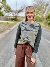 Woman wearing a camouflage sweater and brown pants standing on a road.
