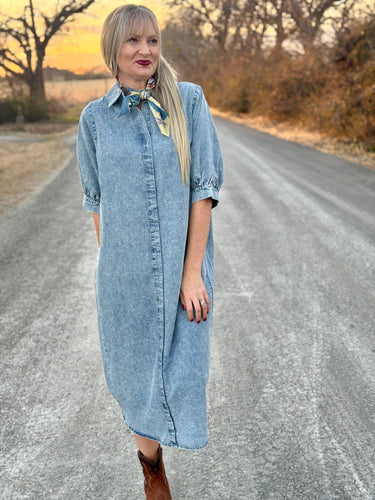 Woman in a denim dress standing on a rural road with trees and sunset in the background