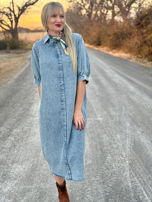 Woman in a denim dress standing on a rural road with trees and sunset in the background