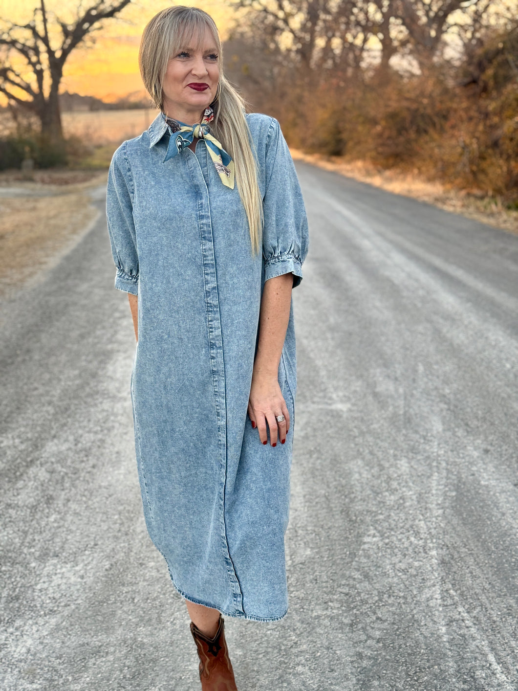 Woman in a denim dress standing on a rural road with trees and sunset in the background
