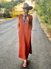 Woman in a rust-colored dress and cowboy hat standing on a road with greenery around.