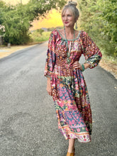 A woman stands on a paved road wearing a long-sleeved bohemian-style midi dress with a highly colorful, busy floral and geometric patchwork print. She is accessorized with turquoise jewelry and brown shoes, with trees and a sunset visible in the background.