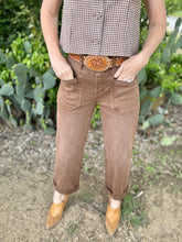 Person wearing brown pants and a checkered shirt with a belt buckle, standing in front of cacti.