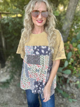 A woman wearing a short-sleeve patchwork t-shirt with a solid mustard yellow upper section and a multicolor floral patchwork lower section, paired with blue jeans and sunglasses, standing outdoors in front of a blurred natural background.
