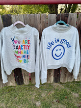 Two gray sweatshirts with motivational text and a smiley face graphic hanging on a wooden fence.