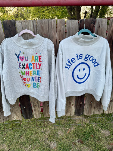 Two gray sweatshirts with motivational text and a smiley face graphic hanging on a wooden fence.