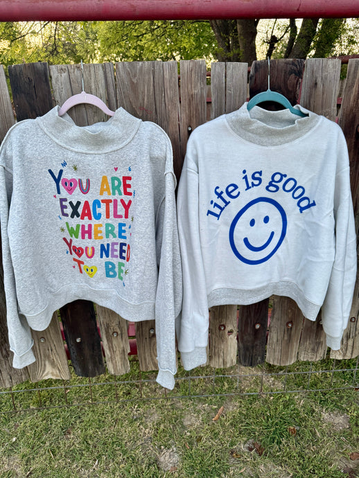 Two gray sweatshirts with motivational text and a smiley face graphic hanging on a wooden fence.
