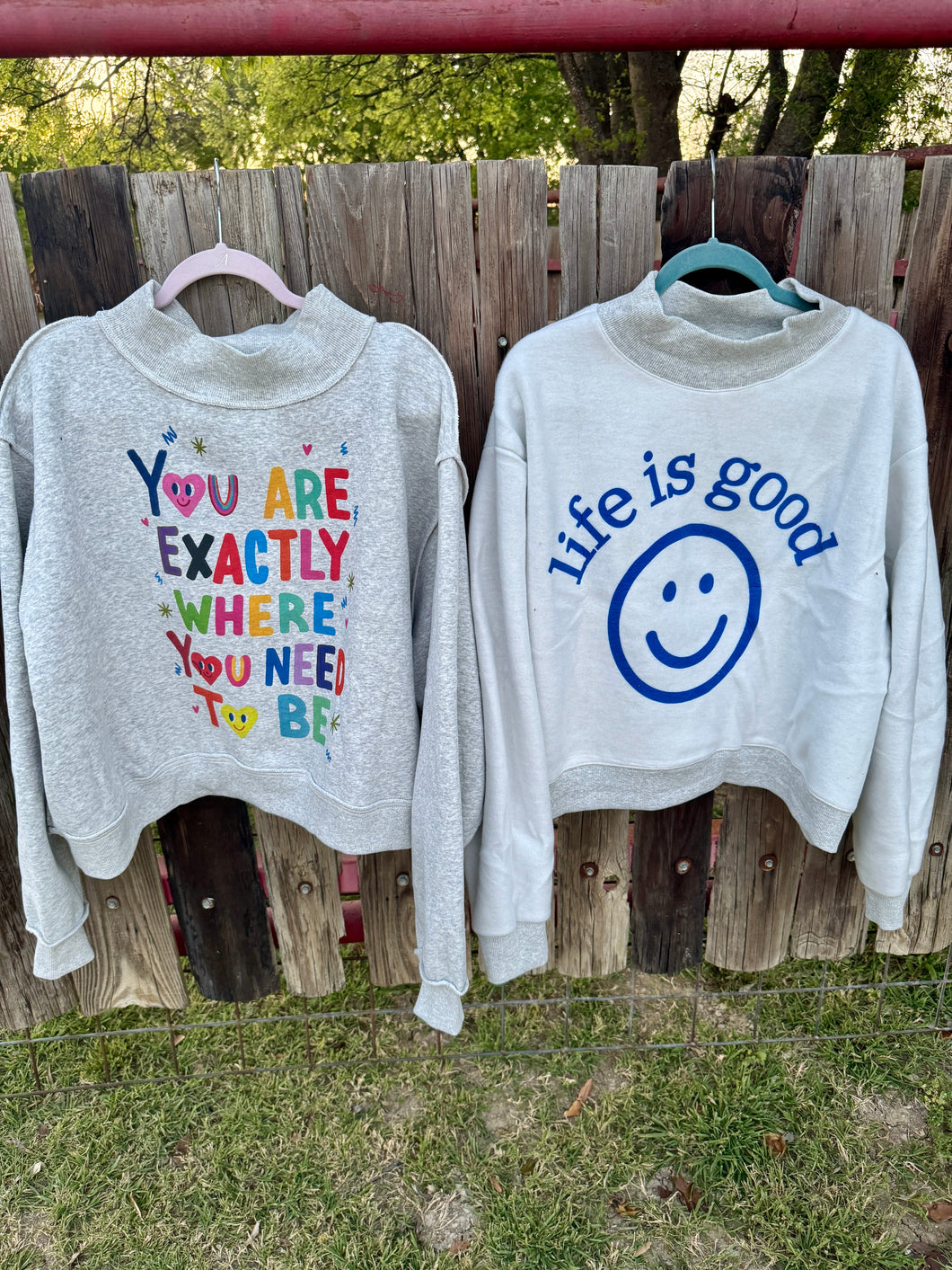 Two gray sweatshirts with motivational text and a smiley face graphic hanging on a wooden fence.