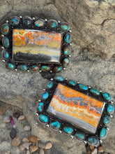 Two rectangular silver-toned belt buckles pieces with turquoise-blue stones and orange marbled centers, displayed on a natural rock surface.