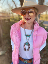 Person wearing a pink puffer vest, white shirt, blue jeans, and a wide-brimmed hat with sunglasses.