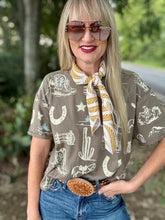 Woman wearing a patterned shirt with cowboy graphics, sunglasses, and a scarf outdoors.