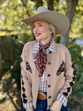 Woman wearing a beige cardigan with geometric patterns, a plaid shirt, and a wide-brimmed hat outdoors.