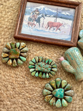 Three turquoise and brown stone flower-shaped belt buckles arranged on a woven tan mat, with a small framed western painting and a cactus-shaped object in the background.