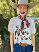 Woman wearing a cowboy hat, scarf, and 'Wild West' t-shirt outdoors.