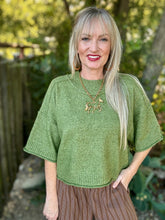 A woman with blonde hair wearing a green short-sleeve knit top and brown pinstriped pants, standing outdoors in front of a blurred garden background.