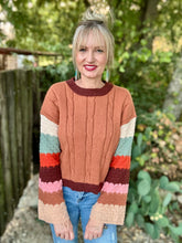 A woman wearing a retro-style cable-knit sweater with a terracotta orange body and multicolor striped sleeves, standing outdoors in front of a wooden fence and green foliage.