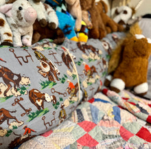 A close-up of a gray bolster pillow with a western cow and rider print, styled on a bed with a colorful patchwork quilt and plush toys in the background.