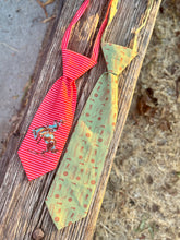 Two novelty neckties displayed on a weathered wooden board outdoors. The tie on the left is red and white striped with a cowboy riding a bucking horse graphic.