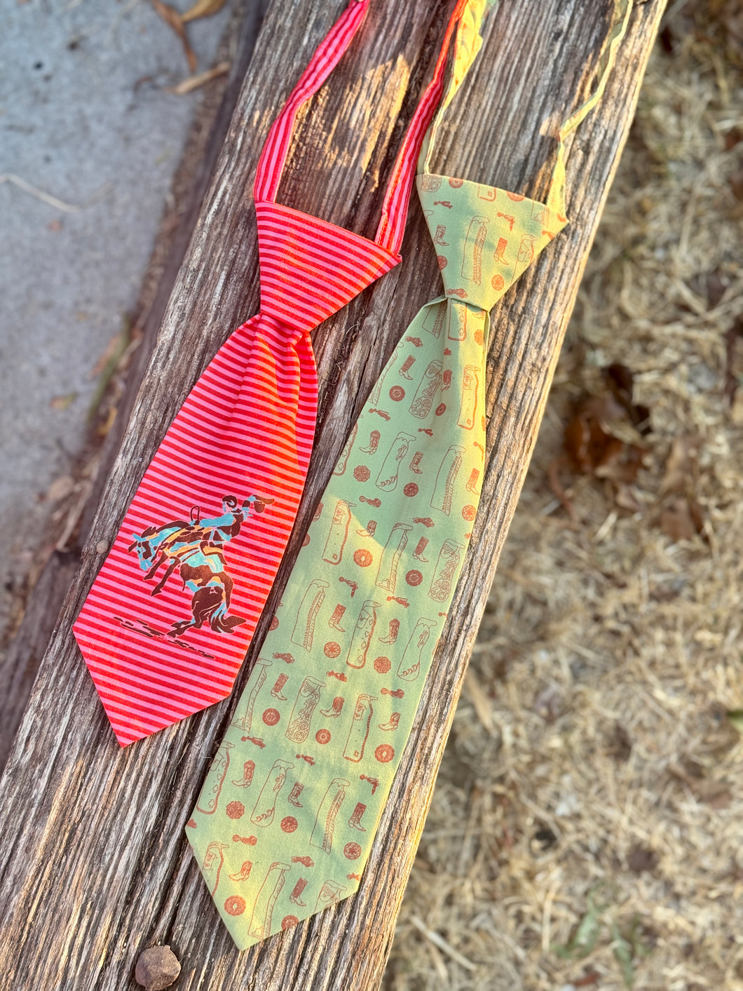 Two novelty neckties displayed on a weathered wooden board outdoors. The tie on the left is red and white striped with a cowboy riding a bucking horse graphic.