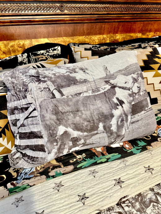 A western-themed bedding set featuring a printed pillow with a black and white cow photograph, a patterned quilt with star motifs, and matching patterned pillows in front of a dark wood headboard.