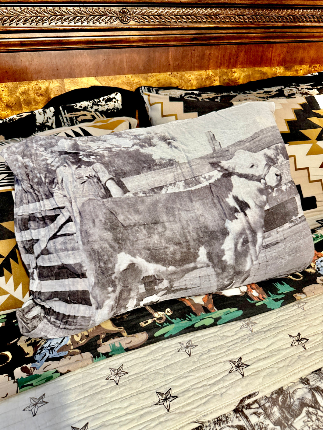 A western-themed bedding set featuring a printed pillow with a black and white cow photograph, a patterned quilt with star motifs, and matching patterned pillows in front of a dark wood headboard.