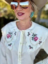 Woman wearing a white blouse with floral embroidery, sunglasses, and a wide-brimmed hat.