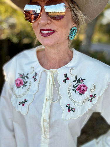 Woman wearing a white blouse with floral embroidery, sunglasses, and a wide-brimmed hat.