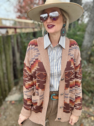 Person wearing a patterned cardigan, sunglasses, and a hat outdoors.