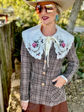 Person wearing a plaid jacket with floral embroidery, sunglasses, and a cowboy hat outdoors.
