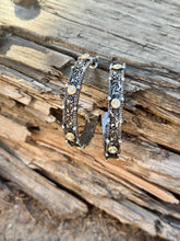 A pair of ornate silver-toned hoop earrings with intricate filigree detailing and small gold-toned circular accents, displayed on a weathered piece of wood outdoors.