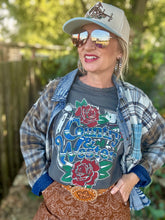 A woman wearing a gray graphic t-shirt with 'Country Western' and rose designs, layered under a blue plaid flannel shirt. She is also wearing a beige baseball cap, blue-tinted sunglasses, and a brown paisley skirt, posed outdoors against a blurred background of trees and a fence.