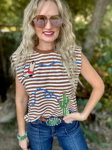 A woman wearing a brown and white striped sleeveless top with embroidered cactus and desert designs, paired with blue jeans and sunglasses, standing outdoors against a blurred green background.