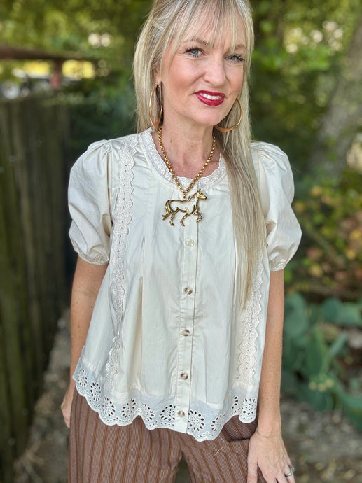 A woman wearing a cream-colored short-sleeve blouse with lace trim and a gold horse pendant necklace, standing outdoors in front of a blurred fence and greenery.