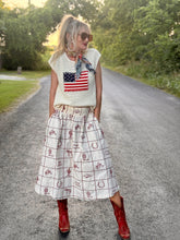 Woman wearing a white sleeveless top with an American flag design and a patterned skirt on a road.