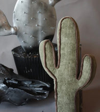 A cactus-shaped ribbed fabric cushion in sage green with brown piping, styled in a rustic arrangement with a metallic cactus and a black skull on a neutral background.