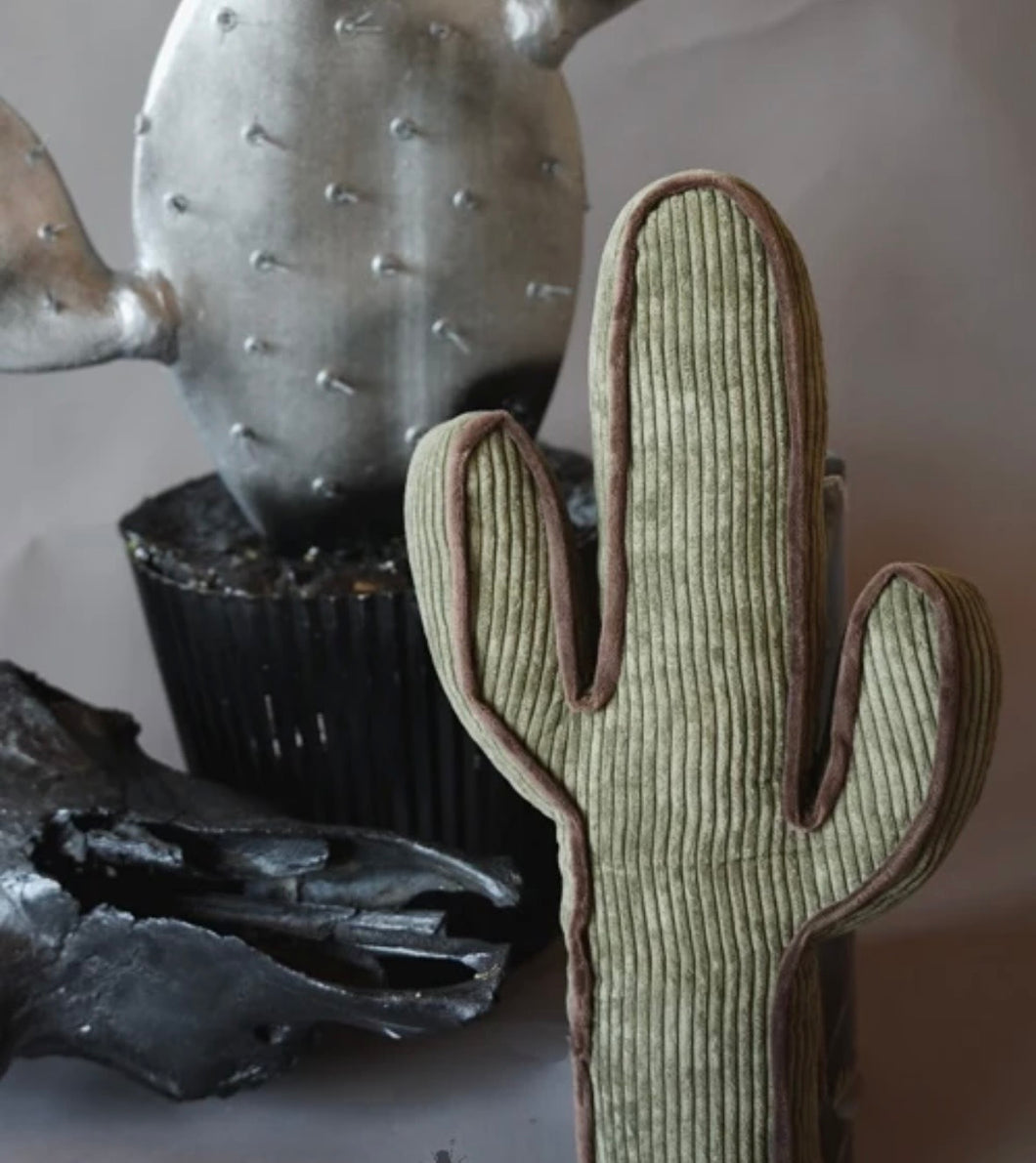 A cactus-shaped ribbed fabric cushion in sage green with brown piping, styled in a rustic arrangement with a metallic cactus and a black skull on a neutral background.