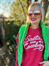 Person wearing a red shirt with 'Santa, bring me a cowboy' text, green cardigan, and sunglasses outdoors.