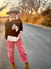 Person wearing a sweater with a horse design, faded red pants, and a hat on a road.