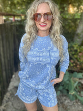 A woman with long blonde wavy hair and sunglasses wearing a light blue and white paisley bandana print long-sleeve top and matching shorts, standing outdoors in front of a wooden fence and greenery.