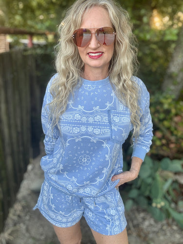 A woman with long blonde wavy hair and sunglasses wearing a light blue and white paisley bandana print long-sleeve top and matching shorts, standing outdoors in front of a wooden fence and greenery.