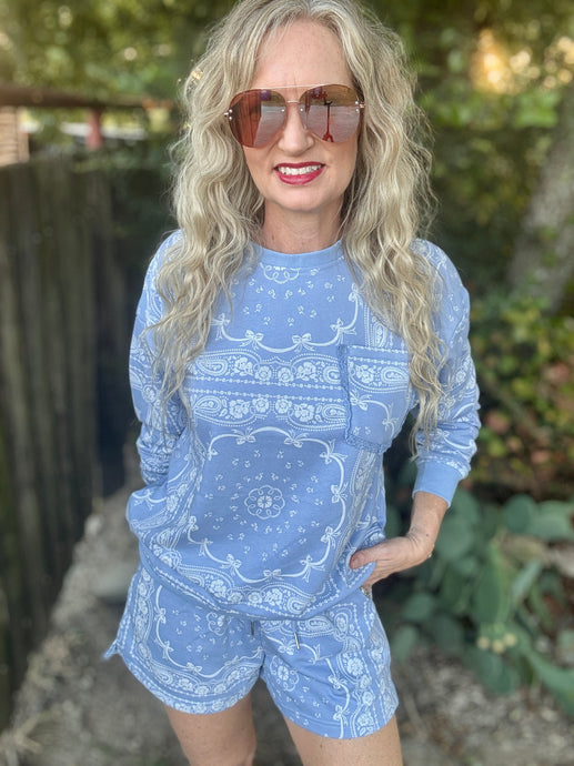 A woman with long blonde wavy hair and sunglasses wearing a light blue and white paisley bandana print long-sleeve top and matching shorts, standing outdoors in front of a wooden fence and greenery.