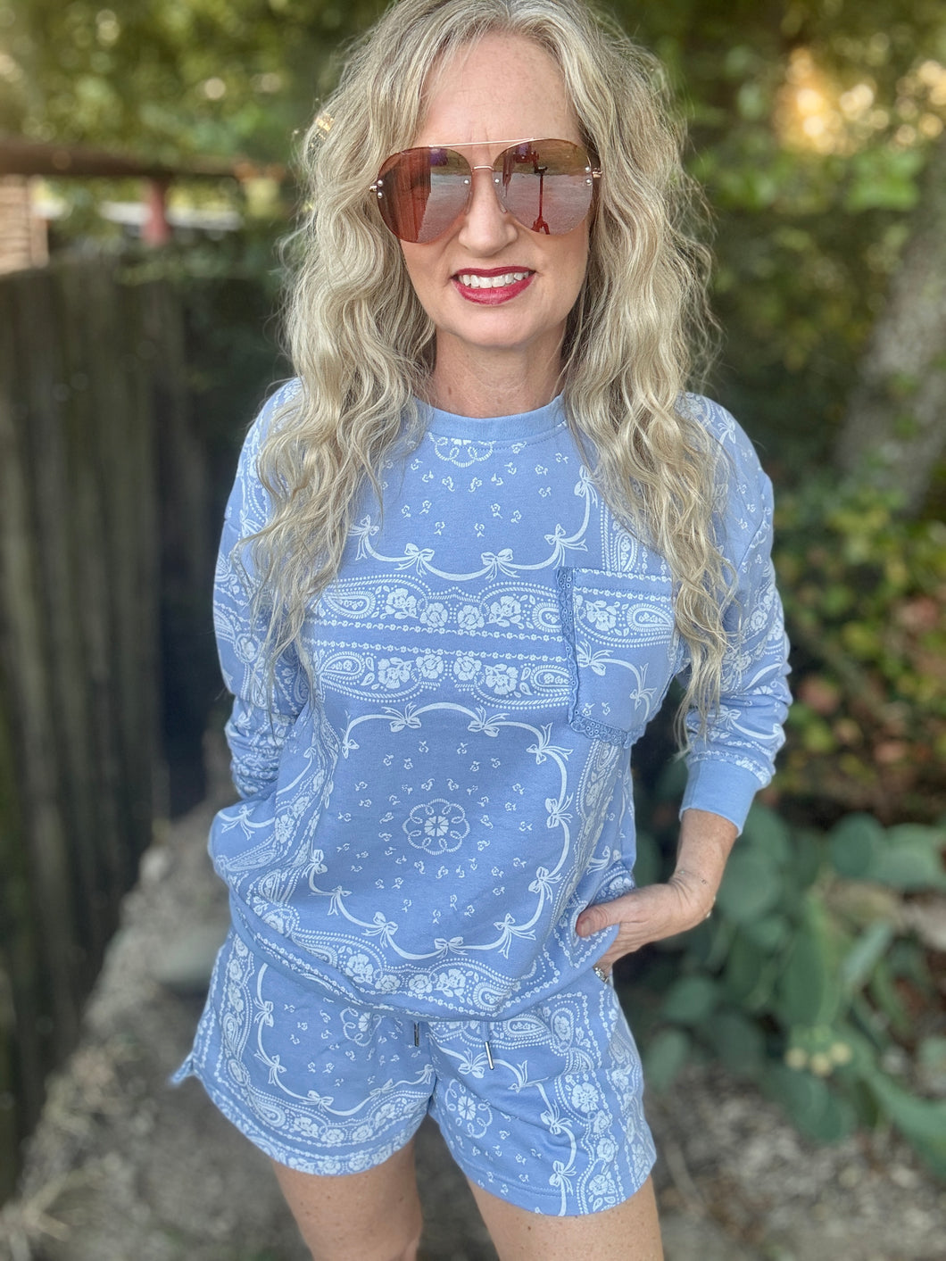 A woman with long blonde wavy hair and sunglasses wearing a light blue and white paisley bandana print long-sleeve top and matching shorts, standing outdoors in front of a wooden fence and greenery.