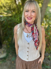 A smiling blonde woman outdoors wearing a cream cable-knit sleeveless vest over a white top, with a red and blue patterned neck scarf and a brown pleated skirt.