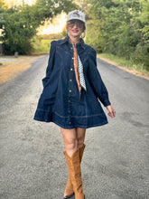 A woman stands on a paved country road wearing a dark blue denim long-sleeve button-up shirt dress, tan cowboy boots, a patterned neck scarf, and a baseball cap.