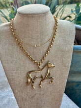 A gold-toned necklace with a horse-shaped pendant displayed on a beige fabric jewelry bust. The necklace has a chunky chain and a delicate layered chain with small crescent moon and star charms.