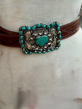 A western-style belt with a large ornate silver-tone buckle featuring turquoise stones and a central turquoise heart, shown on a cream fabric mannequin torso.